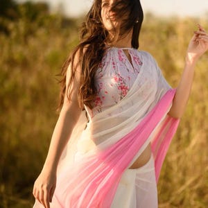 Puede incluir: Un sari blanco con un borde rosa degradado. La blusa tiene un estampado floral en rosa y blanco. El sari está drapeado sobre el cuerpo. El fondo es un campo de hierba alta.