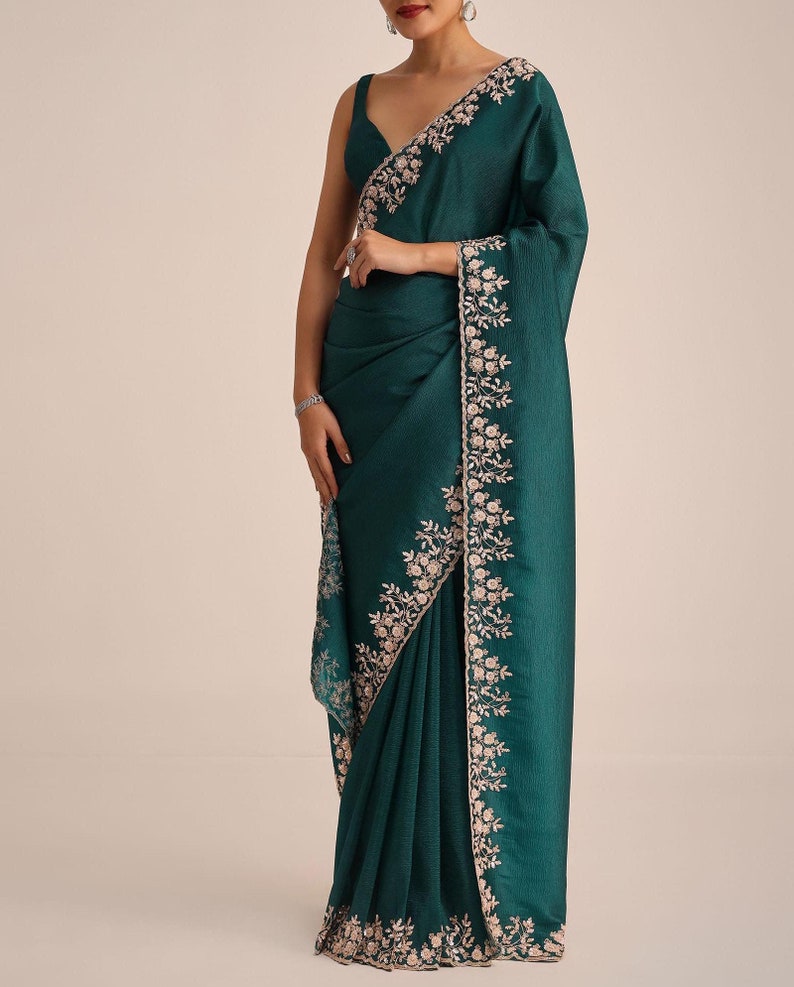May include: A deep emerald green saree featuring a sleeveless blouse and a matching drape. The fabric has a subtle textured finish, and the borders are adorned with intricate light pink floral embroidery and delicate sequin detailing throughout.