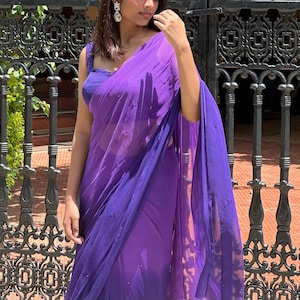 May include: A vibrant purple saree featuring a sheer, lightweight fabric with subtle, scattered embellishments. The garment is paired with a matching sleeveless blouse, creating a cohesive monochromatic look with a flowing drape and elegant texture.