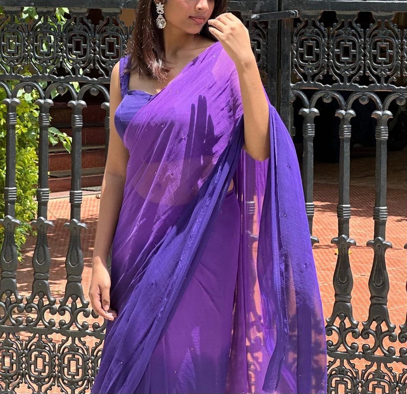 May include: A vibrant purple saree featuring a sheer, lightweight fabric with subtle, scattered embellishments. The garment is paired with a matching sleeveless blouse, creating a cohesive monochromatic look with a flowing drape and elegant texture.
