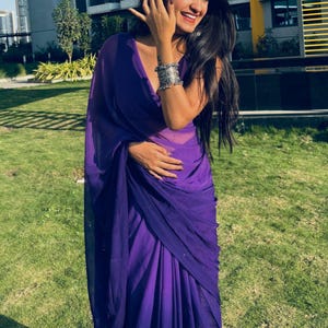 May include: A vibrant purple saree featuring a lightweight, semi-sheer fabric with a smooth drape. The garment is styled with a deep V-neckline and is accessorized with a wide, ornate silver-toned metal cuff bracelet worn on the left wrist.