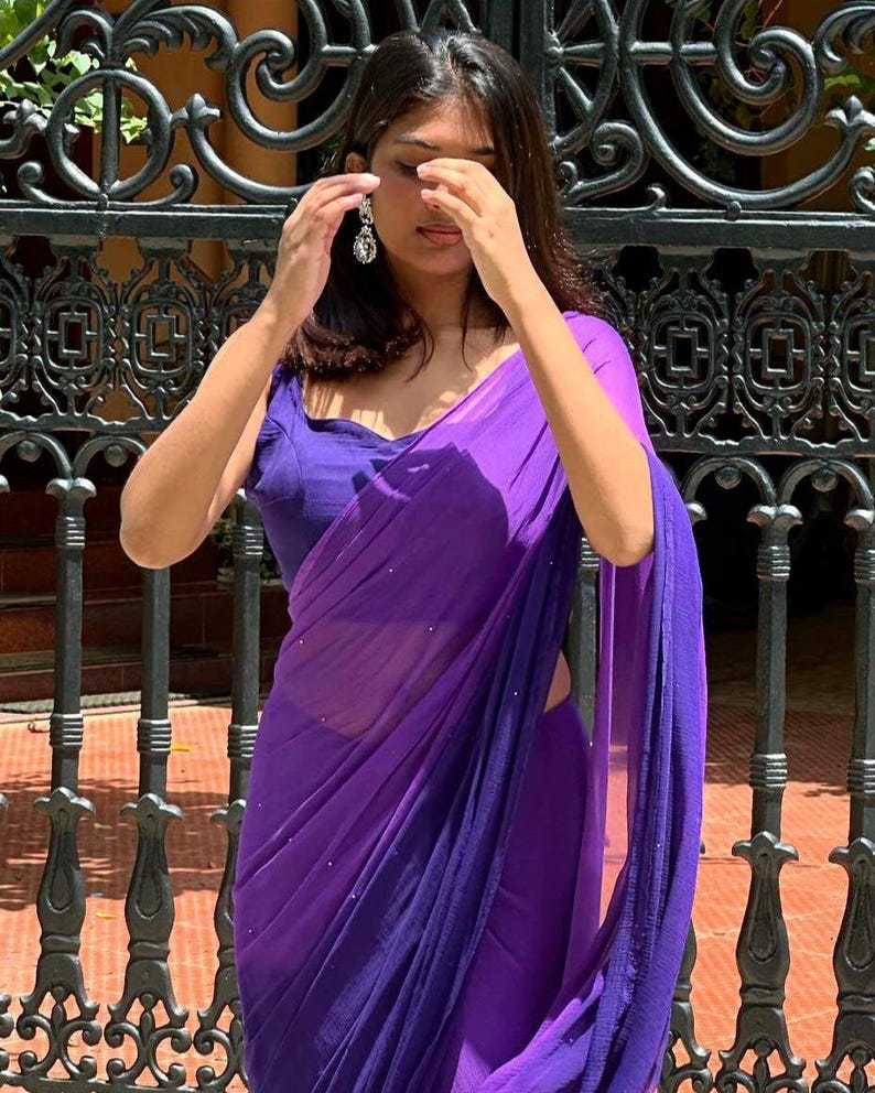 May include: A vibrant purple saree featuring a sleeveless blouse and a sheer, lightweight drape adorned with subtle, scattered silver embellishments. The garment showcases a deep violet hue with a flowing, pleated texture that wraps around the torso.
