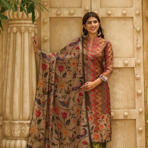 Puede incluir: Una mujer con un salwar kameez indio tradicional. El atuendo presenta una parte superior estampada de manga larga con cuello alto, combinada con pantalones verde oliva. Un chal floral a juego cae sobre los hombros. El fondo incluye un pilar de piedra y una puerta de madera.