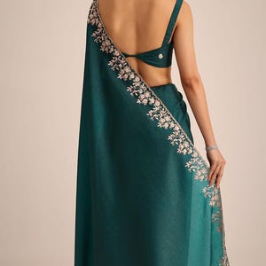 May include: A deep teal saree featuring a sleeveless blouse with a back cutout and a matching skirt. The fabric has a textured finish, and the border is adorned with intricate floral embroidery in light pink and silver metallic thread.