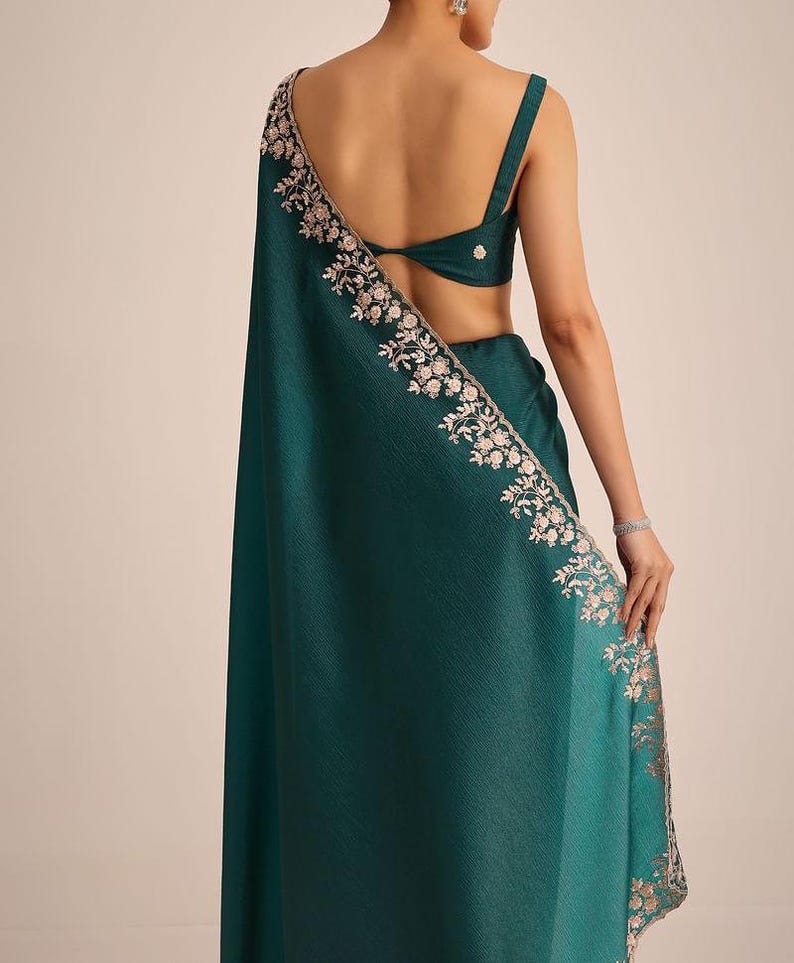May include: A deep teal saree featuring a sleeveless blouse with a back cutout and a matching skirt. The fabric has a textured finish, and the border is adorned with intricate floral embroidery in light pink and silver metallic thread.