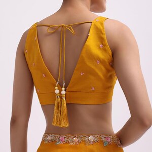May include: A mustard yellow sleeveless blouse featuring a deep V-shaped back with thin tie-up strings ending in decorative gold-toned tassels with white beads. The blouse is adorned with small scattered pink floral embroidery and sequins.