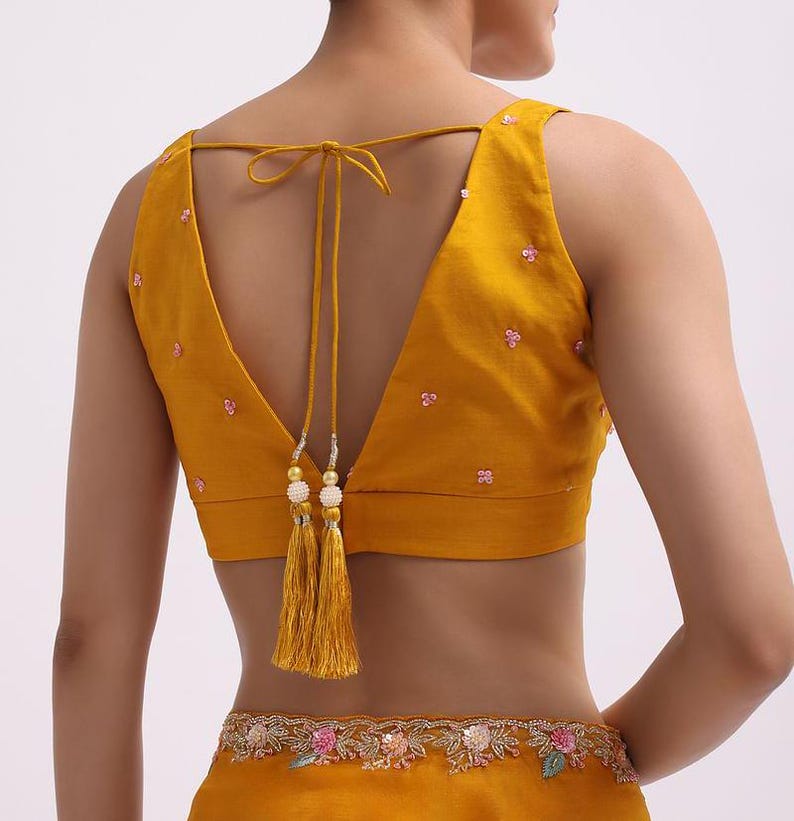 May include: A mustard yellow sleeveless blouse featuring a deep V-shaped back with thin tie-up strings ending in decorative gold-toned tassels with white beads. The blouse is adorned with small scattered pink floral embroidery and sequins.