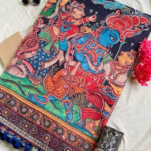 May include: A black fabric textile featuring a vibrant, multi-colored traditional Indian painting of figures with blue skin playing a flute. The border displays intricate floral patterns, and the edge is finished with small blue tassels.