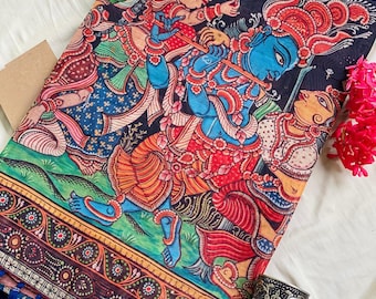 May include: A black fabric textile featuring a vibrant, multi-colored traditional Indian painting of figures with blue skin playing a flute. The border displays intricate floral patterns, and the edge is finished with small blue tassels.
