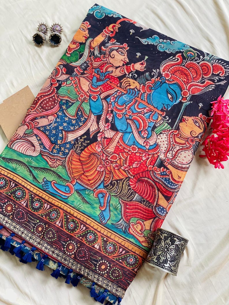 May include: A black fabric textile featuring a vibrant, multi-colored traditional Indian painting of figures with blue skin playing a flute. The border displays intricate floral patterns, and the edge is finished with small blue tassels.