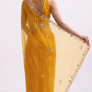 May include: A mustard yellow saree featuring a sheer pallu with delicate floral embroidery along the edges. The garment includes a sleeveless blouse with a V-neckline and thin tie-up straps at the back, adorned with small scattered floral motifs.