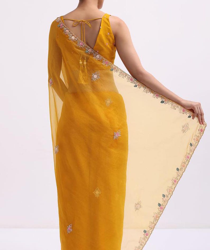 May include: A mustard yellow saree featuring a sheer pallu with delicate floral embroidery along the edges. The garment includes a sleeveless blouse with a V-neckline and thin tie-up straps at the back, adorned with small scattered floral motifs.