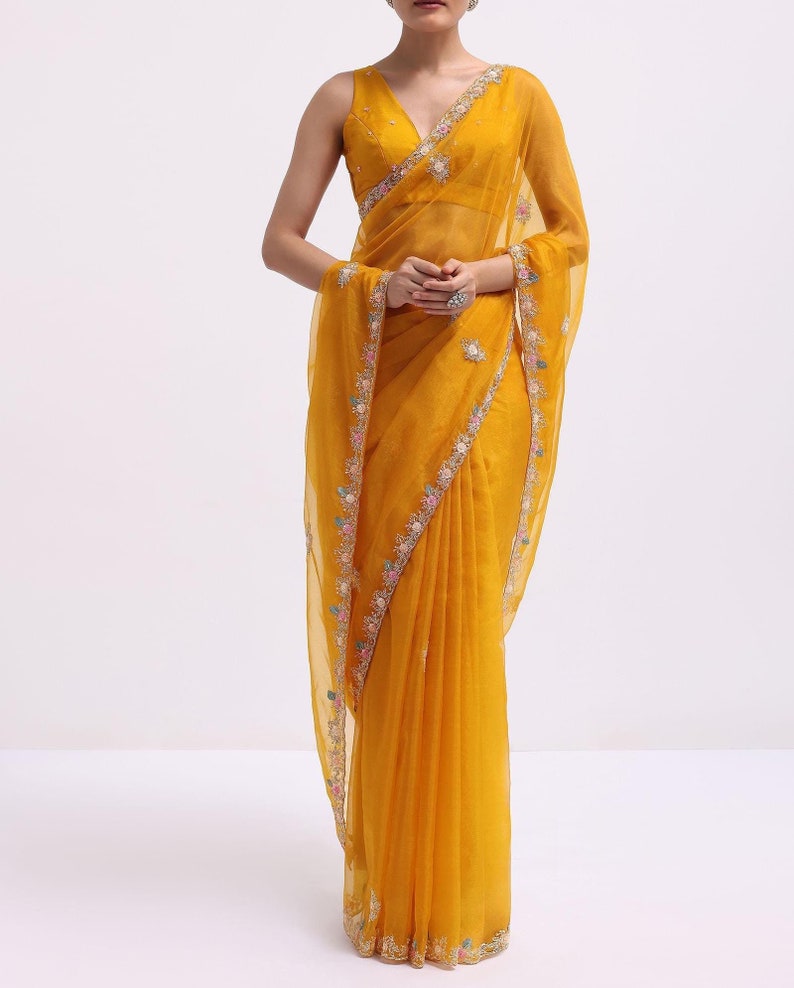 May include: A vibrant mustard yellow saree featuring a sleeveless blouse with a deep V-neckline. The sheer fabric of the saree is adorned with a delicate floral embroidery border in shades of pink, blue, and white, accented with small silver sequins.