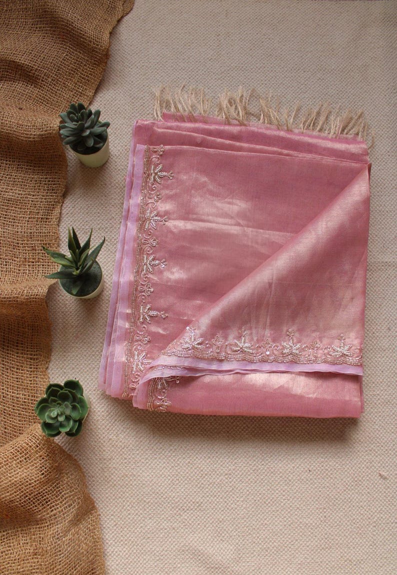 Pu&ograve; includere: Una sciarpa rosa polvere piegata con una lucentezza sottile e un bordo delicatamente ricamato con motivi floreali in filo d'argento. La sciarpa &egrave; rifinita con frange beige chiaro sul bordo superiore, realizzata in un tessuto morbido e leggero.