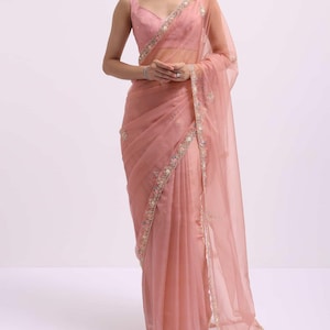 May include: A dusty rose saree featuring a sleeveless blouse and a sheer, lightweight fabric drape. The garment is adorned with intricate, multi-colored floral embroidery along the borders and scattered throughout the fabric, creating a delicate look.
