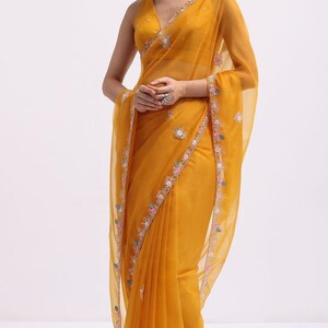 May include: A mustard yellow saree featuring a sheer, lightweight fabric with a V-neck sleeveless blouse. The garment is adorned with intricate floral embroidery along the borders and scattered motifs in shades of pink, blue, and white thread.