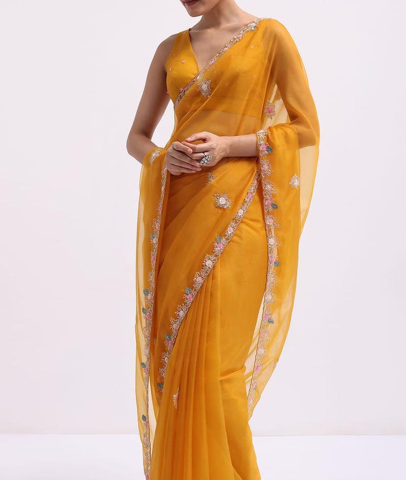 May include: A mustard yellow saree featuring a sheer, lightweight fabric with a V-neck sleeveless blouse. The garment is adorned with intricate floral embroidery along the borders and scattered motifs in shades of pink, blue, and white thread.