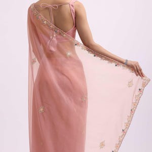 May include: A dusty rose saree featuring a sheer, lightweight fabric with delicate floral embroidery along the borders and scattered across the body. The garment includes a matching sleeveless blouse with a tie-back closure and tassel details.