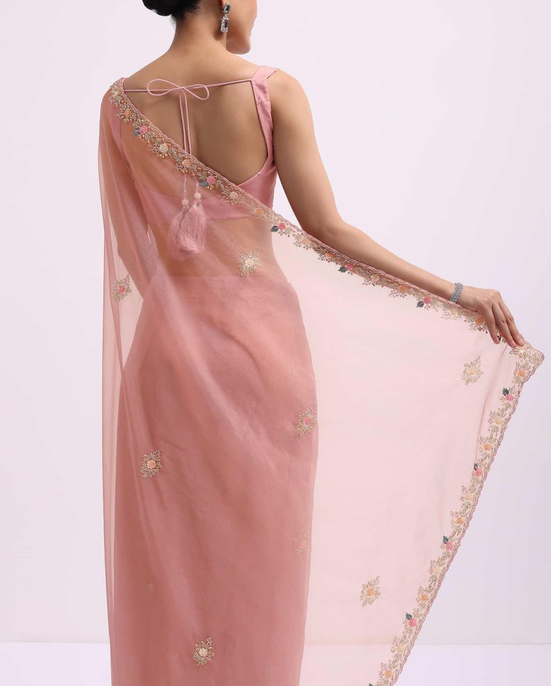 May include: A dusty rose saree featuring a sheer, lightweight fabric with delicate floral embroidery along the borders and scattered across the body. The garment includes a matching sleeveless blouse with a tie-back closure and tassel details.