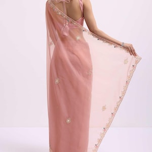 May include: A sheer dusty rose saree featuring delicate floral embroidery along the borders and scattered motifs throughout the fabric. The garment is paired with a matching sleeveless blouse that includes a tie-back closure with tassel details.