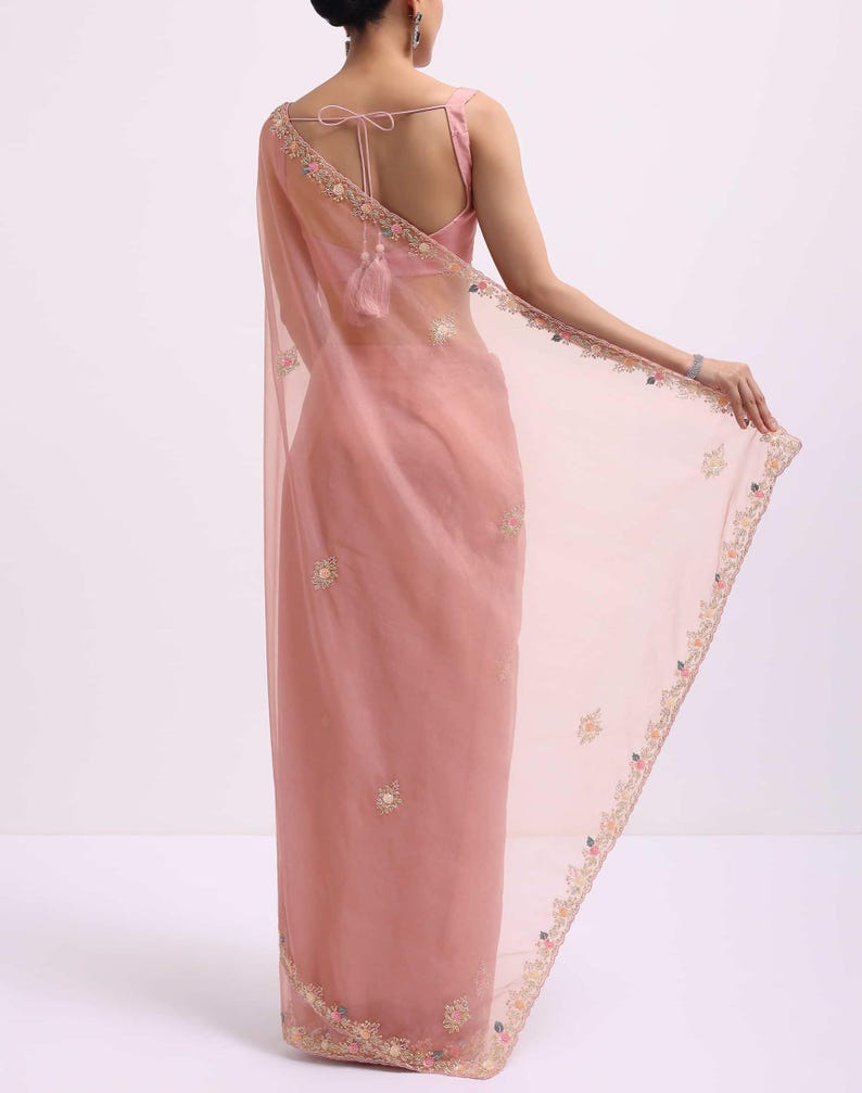 May include: A sheer dusty rose saree featuring delicate floral embroidery along the borders and scattered motifs throughout the fabric. The garment is paired with a matching sleeveless blouse that includes a tie-back closure with tassel details.