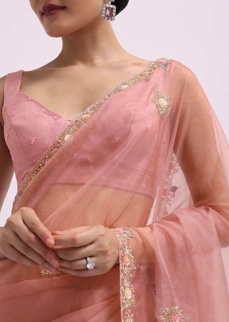 May include: A blush pink saree with detailed floral embroidery on the borders and sleeves. The sheer fabric drapes elegantly, paired with a matching blouse. Delicate embellishments add a touch of glamour to this formal wear.