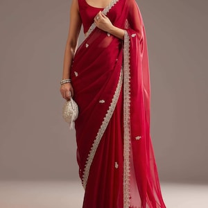 May include: A deep red saree featuring a sheer fabric with intricate silver embroidery along the borders and scattered floral motifs. The garment is paired with a matching sleeveless blouse and a small, round, silver-toned beaded clutch bag.