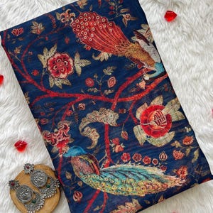 May include: A navy blue fabric with a vibrant peacock and floral design in red, orange, and teal. The fabric is adorned with intricate details, and a pair of silver earrings with green accents rests on a wooden coaster.