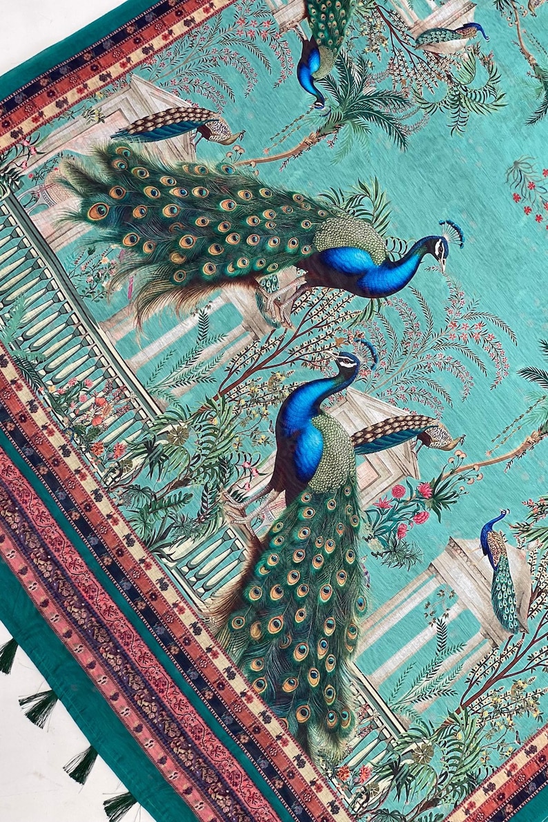 May include: A teal fabric featuring a repeating pattern of peacocks with iridescent blue and green plumage. The design includes architectural structures, palm trees, and floral elements. The edge has a decorative border with tassels.