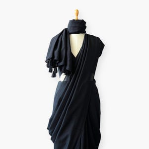 Puede incluir: Un sari negro y una bufanda a juego con borlas exhibidas en un maniquí. El sari está drapeado para mostrar su diseño, con la bufanda elegantemente drapeada alrededor del cuello y los hombros. El fondo es blanco liso.