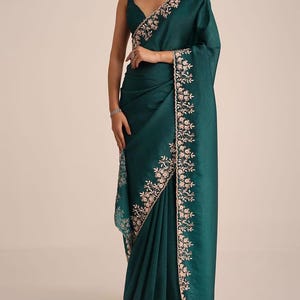 May include: A deep teal saree featuring a sleeveless blouse with a V-neckline. The fabric has a subtle textured finish and is adorned with an intricate floral embroidery border in light pink and white thread along the edges and the pallu.