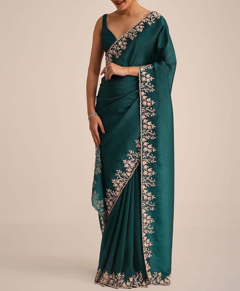 May include: A deep teal saree featuring a sleeveless blouse with a V-neckline. The fabric has a subtle textured finish and is adorned with an intricate floral embroidery border in light pink and white thread along the edges and the pallu.