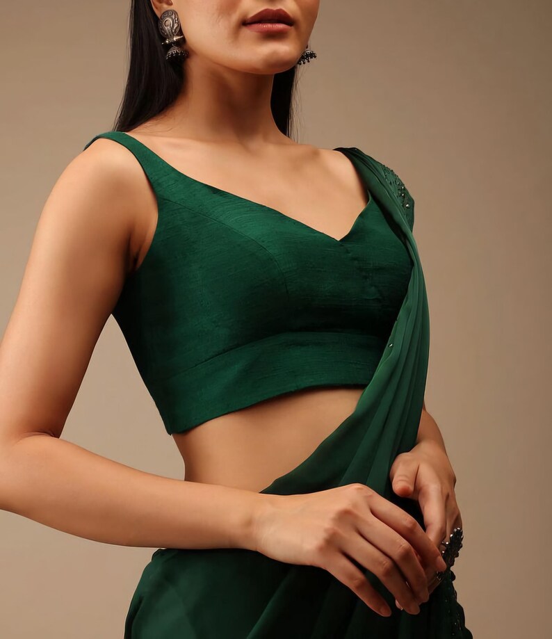 Puede incluir: Una blusa sin mangas de color verde esmeralda oscuro con escote en V y una cintura ancha, combinada con una prenda de tela drapeada a juego en verde esmeralda oscuro con sutiles adornos de cuentas a lo largo del borde.