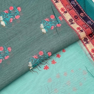 May include: A teal fabric featuring a repeating pattern of embroidered pink and white floral sprigs. The fabric includes a decorative border with horizontal stripes in shades of red, cream, and dark blue, accented with small floral motifs.