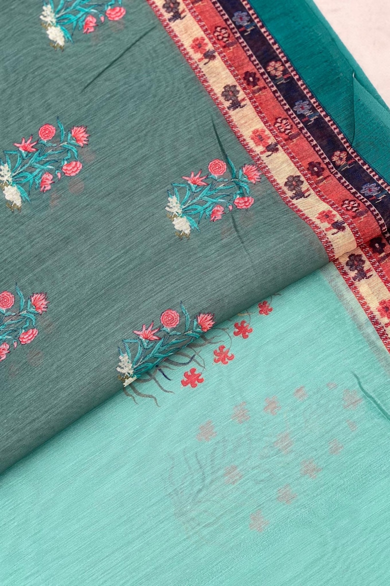 May include: A teal fabric featuring a repeating pattern of embroidered pink and white floral sprigs. The fabric includes a decorative border with horizontal stripes in shades of red, cream, and dark blue, accented with small floral motifs.