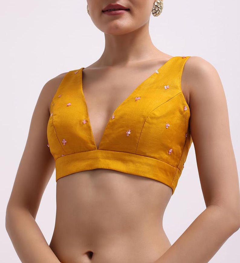 May include: A mustard yellow sleeveless blouse featuring a deep V-neckline and a cropped hem. The fabric is adorned with small, scattered pink floral-shaped sequin embellishments across the front panels, providing a subtle texture and shine.