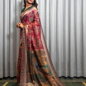 May include: A red and brown silk saree with a peacock and floral print. The saree has a gold border with intricate designs. The saree is draped on a woman standing in front of a white curtain.