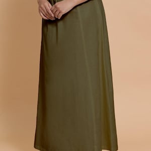 May include: Olive green maxi skirt with a drawstring waist. The skirt is made of a flowing fabric and falls to the ankles. The model is wearing brown embroidered flats. The background is a solid tan color.
