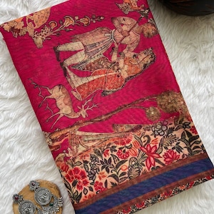 May include: A vibrant magenta fabric featuring a detailed print of two figures in traditional attire, a deer, and floral motifs. The bottom border displays a dark blue stripe with intricate botanical patterns. Two silver earrings are placed nearby.