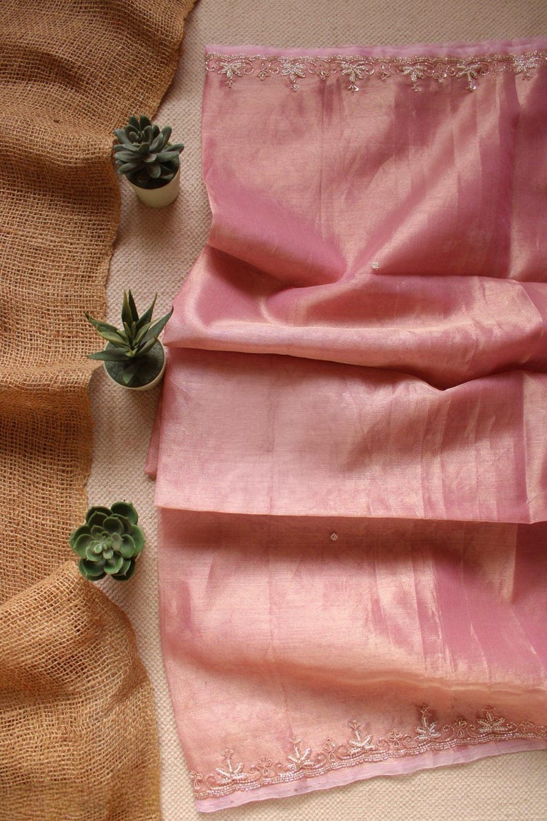 Pu&ograve; includere: Un tessuto rosa chiaro con una lucentezza sottile e una consistenza semi-trasparente. Il tessuto presenta un ricamo delicato e complesso lungo i bordi superiore e inferiore, composto da piccoli motivi floreali accentuati da minuscole perline argentate.