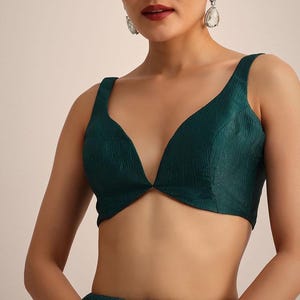 May include: A sleeveless, deep V-neck blouse in a dark emerald green color. The fabric features a subtle, crinkled, textured finish throughout. The garment has wide shoulder straps and a fitted silhouette that ends just above the waistline.