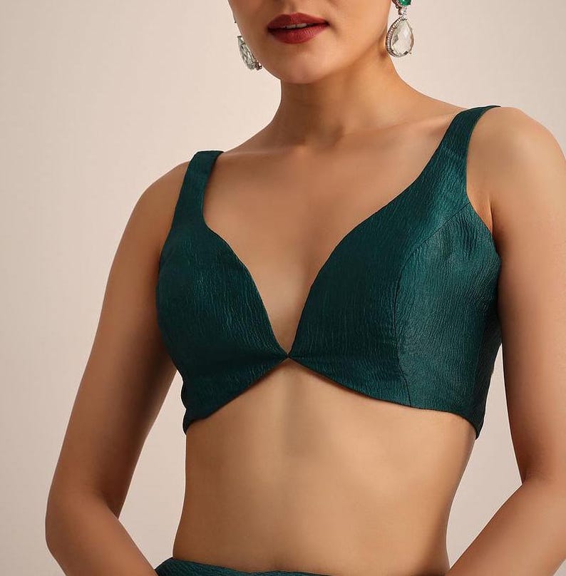 May include: A sleeveless, deep V-neck blouse in a dark emerald green color. The fabric features a subtle, crinkled, textured finish throughout. The garment has wide shoulder straps and a fitted silhouette that ends just above the waistline.