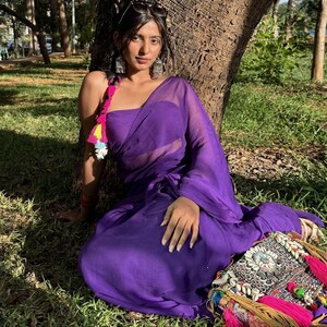 May include: A vibrant purple saree featuring a sheer, lightweight fabric draped over a matching strapless bodice. The ensemble is accented by a colorful braided hair accessory with pink and yellow tassels and a decorative bag with shells and beads.