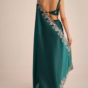 May include: A deep teal sari featuring a sleeveless blouse with a back cutout and a matching drape. The fabric has a subtle texture and is adorned with intricate light-colored floral embroidery along the edges of the long, flowing garment.