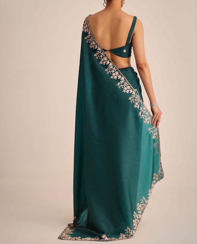 May include: A deep teal sari featuring a sleeveless blouse with a back cutout and a matching drape. The fabric has a subtle texture and is adorned with intricate light-colored floral embroidery along the edges of the long, flowing garment.