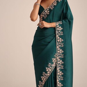 May include: A deep emerald green saree featuring a textured fabric finish and a wide border adorned with intricate light pink floral embroidery. The garment is draped with pleats at the waist and a pallu that falls over the shoulder, showcasing detailed needlework along the edges.