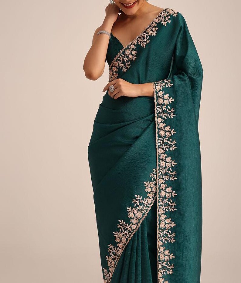 May include: A deep emerald green saree featuring a textured fabric finish and a wide border adorned with intricate light pink floral embroidery. The garment is draped with pleats at the waist and a pallu that falls over the shoulder, showcasing detailed needlework along the edges.