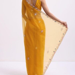 May include: A mustard yellow saree featuring a sheer, lightweight fabric with intricate floral embroidery along the borders and scattered motifs. The blouse has a deep V-neckline with thin tie-up straps at the back and decorative tassels.