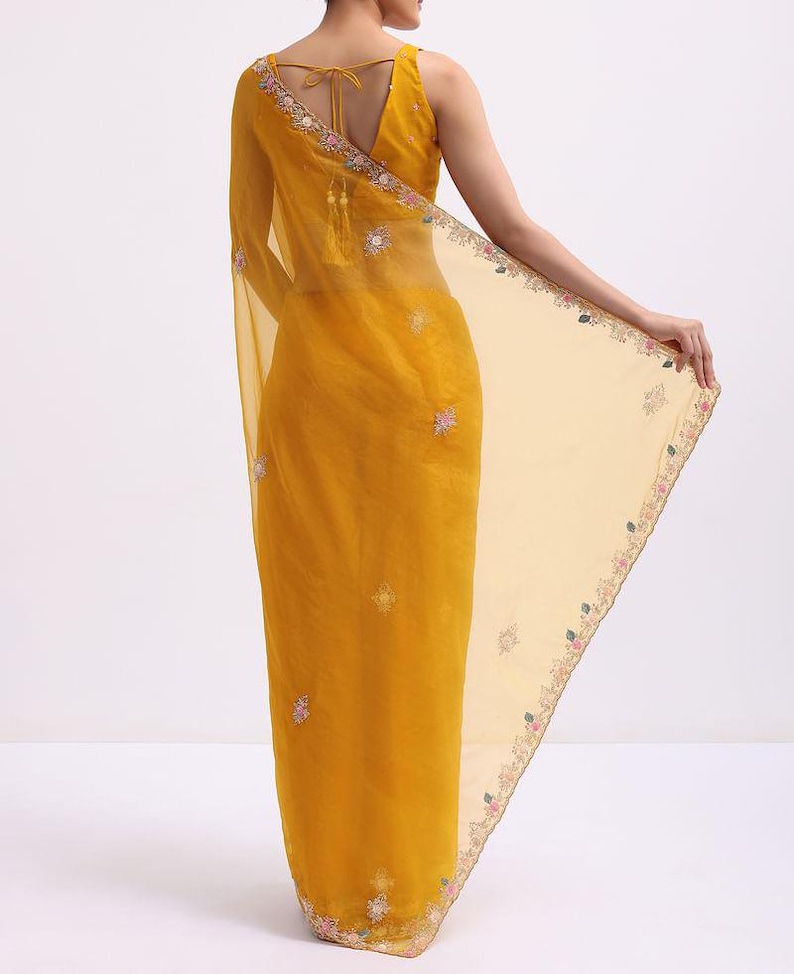 May include: A mustard yellow saree featuring a sheer, lightweight fabric with intricate floral embroidery along the borders and scattered motifs. The blouse has a deep V-neckline with thin tie-up straps at the back and decorative tassels.