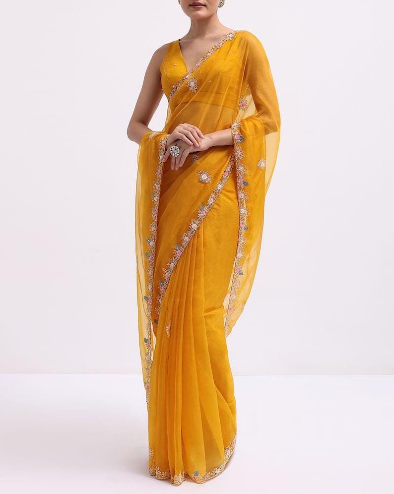 May include: A vibrant mustard yellow saree featuring a sheer, lightweight fabric with a deep V-neck sleeveless blouse. The borders are adorned with intricate floral embroidery in shades of pink, blue, and white, complemented by matching motifs.